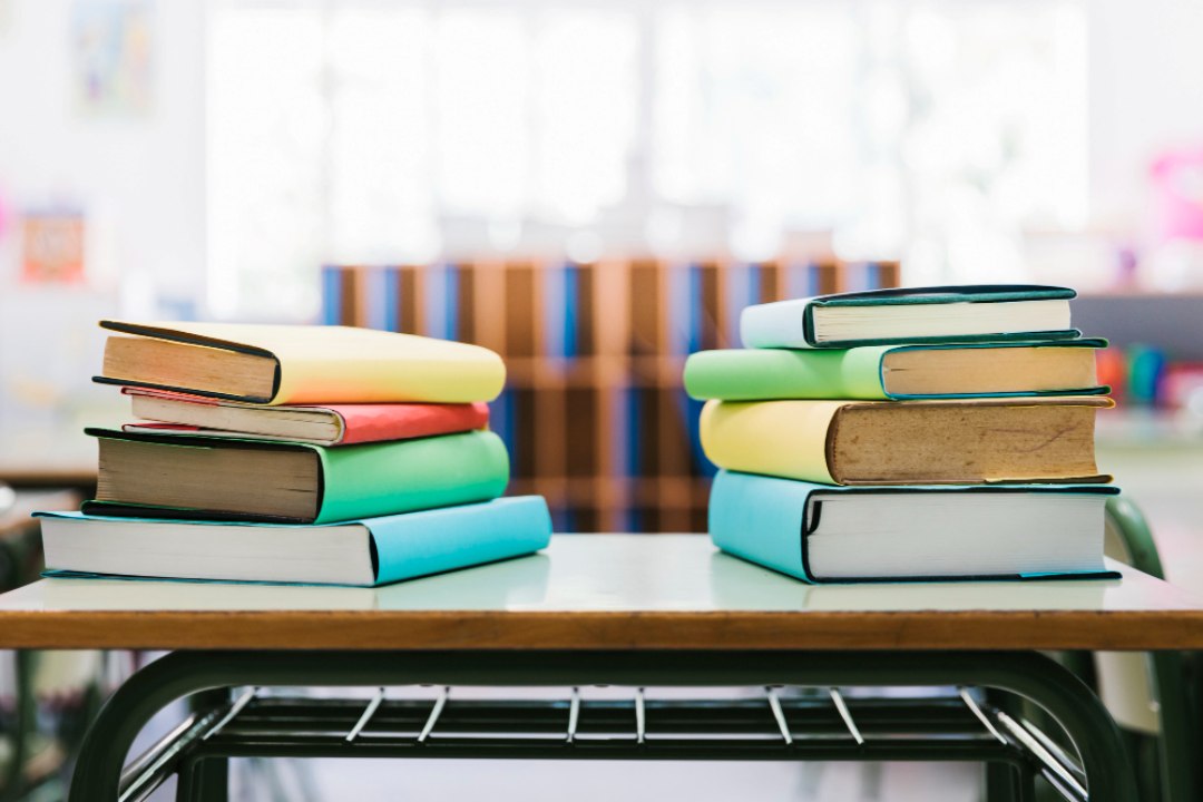 Tumpukan dua set buku teks berwarna di atas meja sekolah mewakili peranan pembekal buku teks dalam menyediakan bahan pembelajaran yang berkualiti.